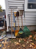 Wide view of 7 garden tools including snow shovel, 2 shovels, 2 green plastic leaf rakes, metal garden rake and broom, leaning against house exterior with fallen leaves at base.