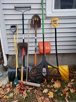 Photo showing seven outdoor garden tools arranged upright against a wall with fallen leaves on the ground. Two plastic snow shovels, two metal rakes, and three metal shovels with wooden or plastic handles visible.
