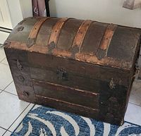 Front and side view of a vintage steamer trunk showing wood slats on top and metal corner protectors. The trunk is resting on a blue patterned rug.