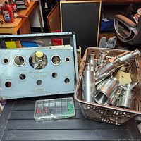 Photo of metal control panel housing, basket with metallic cylindrical parts and a plastic organizer with small parts on a black table.