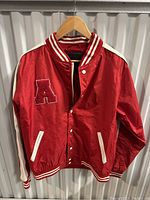 Front view of red satin varsity jacket with white chenille A patch, snap button closure, and white-trimmed pockets on hanger.