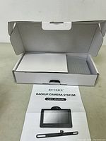 White box partially open with user manual laid flat in front showing backup camera system on cover.