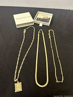 Three gold-tone jewelry necklaces displayed on dark surface with Kissyan branded box and card in background, showing pendant with letter K and two additional chains.