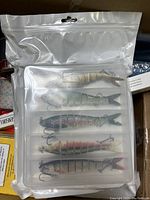 Set of five multi-jointed fish-shaped fishing lures inside clear plastic container in resealable plastic bag.