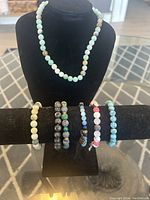 Photo of a natural stone bead necklace displayed on a bust, and six assorted beaded bracelets displayed on a horizontal bar in front of it, showing color and bead details for all jewelry in the set.