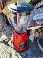 Red Oster blender with glass pitcher and black lid shown on outdoor surface among miscellaneous items.