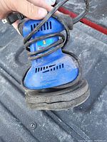 Photo of blue handheld electric sander showing ventilation slots, circular sanding pad, and attached power cord.