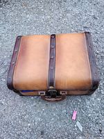 Closed vintage brown leather case on rough ground surface, showing handle and outer structure with metal rivets