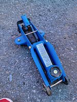 Blue hydraulic floor car jack resting on asphalt surface outdoors. Visible spring and wheels on the jack.