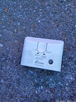 Front view of a rectangular white Kidde carbon monoxide alarm placed on textured ground.