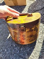 Hand-held photo of vintage carry container showing the oval shape, wooden handle, metal clasp, and exterior with vintage designs.