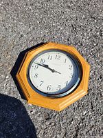 Seiko octagonal wooden wall clock with a white face, black Arabic numerals, black hour and minute hands, brass second hand, and glass front on an asphalt background.