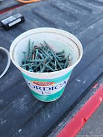 Plastic yogurt container filled with green wood screws on a dark textured surface, some screws pointing up, others lying flat.