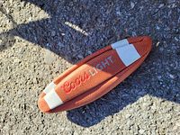 Photo of a small, orange-brown football with white stripes and Coors Light logo embroidered in red, placed on textured ground outdoors.