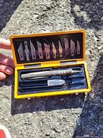 Compact yellow plastic case containing a black and silver cutting tool handle and a variety of ten small metal cutting blades of various shapes and sizes.