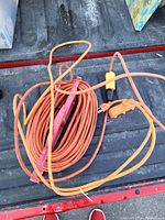 Photo shows three orange extension cords including one coiled with pink strap, laying on a textured black surface with some wear marks.