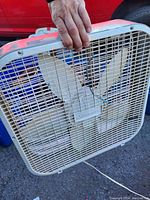 White plastic box fan with three blades being held by a hand in front of a red vehicle.