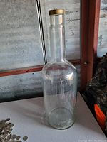 Clear glass jug standing on a white surface with a gold-colored cap. Visible transparency of the glass allows a view through it, showing its cylindrical shape and narrow neck.