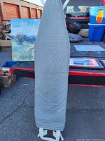 Full image of ironing board with grey cover and white metal legs leaning against a truck bed outdoors.