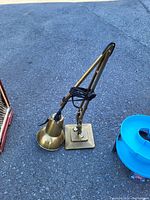 Photo of an adjustable desk lamp with brass finish placed outdoors on concrete. Lamp has a square base, adjustable metal arm, and cone-shaped lamp head. Black electrical cord wrapped around arm.
