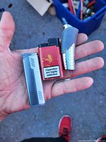 Hand holding three lighters: a slim silver butane lighter, a red and black disposable cigarette lighter with health warning label, and a textured silver torch flame lighter with curved design.