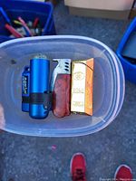 Top-down photo of a plastic container holding three lighters: a blue cylindrical torch lighter with a clip, a metal lighter with brass and gold tones, and a gold-colored disposable lighter in original packaging.