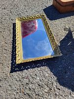 Photo of rectangular mirror with ornate gold frame resting on concrete surface, showing the entire mirror and frame detail.