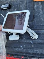 White rectangular solar panel with mounting bracket and attached white cable, along with a black power adapter placed on a dark textured surface.