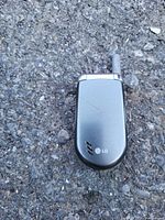 Closed view of an older silver-gray LG flip phone with external antenna, placed on asphalt surface.