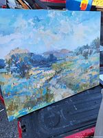 Photo of a landscape painting showing a river, trees, and sky with clouds, placed upright outdoors.