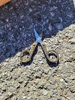 Photo showing small metal scissors with ornate brass-colored handles on asphalt surface.