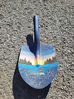Photo of a metal shovel spade laid on asphalt, blade decorated with a colorful hand-painted landscape scene including trees, water, and sky at sunset.