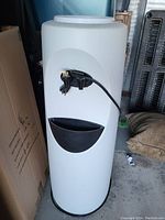 White standing water dispenser with front spout and drip tray shown from front view, surrounded by boxes and other items.