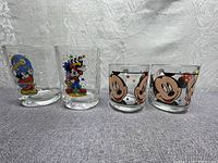 Four clear glasses featuring Walt Disney Mickey Mouse characters, shown side by side on fabric background.