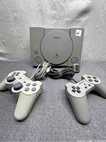 Sony PlayStation 1 SCPH-5501 console with 2 wired controllers. Console and controllers shown from front, cables coiled and in front.