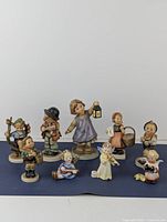 Full view of ten Hummel Goebel figurines featuring children and angels posed with various objects on dark surface in front of white background