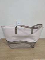 Front view of the two-tone tote bag showing cream upper area and taupe bottom, with some wrinkling on the bottom part, sitting on a wooden surface against a white wall.