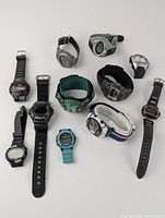 Overview photo showing all 11 digital watches arranged on white surface, various strap materials and colors visible, watches have dead batteries and show wear.