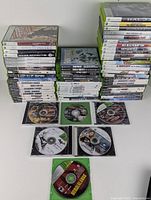 Photo showing stacks of Xbox, Xbox 360, and Xbox One game cases, with some discs laid out in front. Titles include Mass Effect 2, Rainbow Six, BioShock, Halo, Fortnite, Call of Duty, Borderlands, and more.