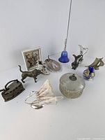 Photo showing entire lot items arranged on white surface including brass iron, dog and boat figurines, glass bell, ceramic boot and pitcher, framed picture, napkin holder, trinket box, and metal plaque.