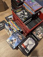 Photo showing a red plastic crate filled with stacked DVD cases including 'Swordfish', 'August Rush', 'Mad Men', and 'M*A*S*H' titles and other assorted DVDs spread out on the floor.
