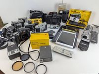 Overview of lot showing vintage cameras, accessories, electronic devices including tablets and smartphones, and remote controls laid out on table.