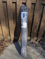 Patio umbrella packed inside a grey fabric carry bag standing upright against a wood fence outdoors on a sunny day.