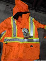 Full view of orange mid-length jacket with reflective yellow and silver stripes, hood, front pockets, and tags attached.