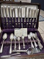 Vintage silver plated flatware set arranged in purple velvet lined case showing knives in upper section, various forks, spoons, ladle, and other utensils in lower section, with some patina and wear.