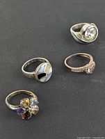 Four sterling silver rings displayed on dark surface, showing various designs including cluster flower with multi-colored stones, pastel colored inlay, a clear stone on rose-gold tone band, and a large clear stone with patina and slight bend.