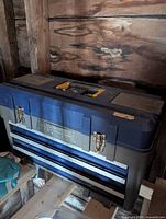 Photo of a dark blue and gray plastic toolbox with two front drawers, metal latch locks, and a yellow handle on top, placed in a wood-paneled area.