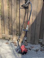 Full view of gasoline-powered weed whacker leaning against a wooden fence showing red and black motor housing, curved metal shaft, ergonomic handle with front handle, and plastic protective guard. Overall outdoor-used condition.