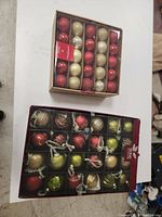 Photo of two boxed sets of Christmas ornaments, one containing round matte and glitter balls in red, gold, and silver, the other containing assorted shaped ornaments including balls and other festive shapes in red, gold, green, and silver.