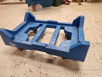 Angled front view of blue plastic bin dolly on a tiled floor, showing raised edge supports and platform surface details.